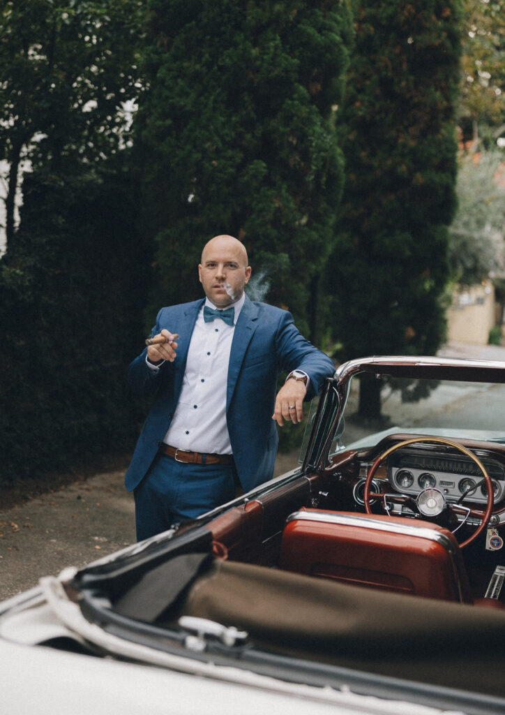 A groom smokes a cigar by the classic car he rented for his Charleston, SC wedding.